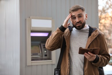 Photo of Unhappy young man with wallet near cash machine outdoors
