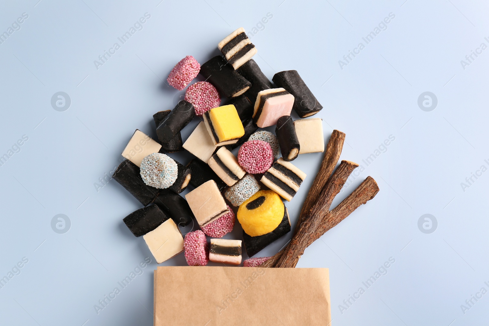 Photo of Paper bag with tasty candies and dried cut liquorice root on light blue background, top view