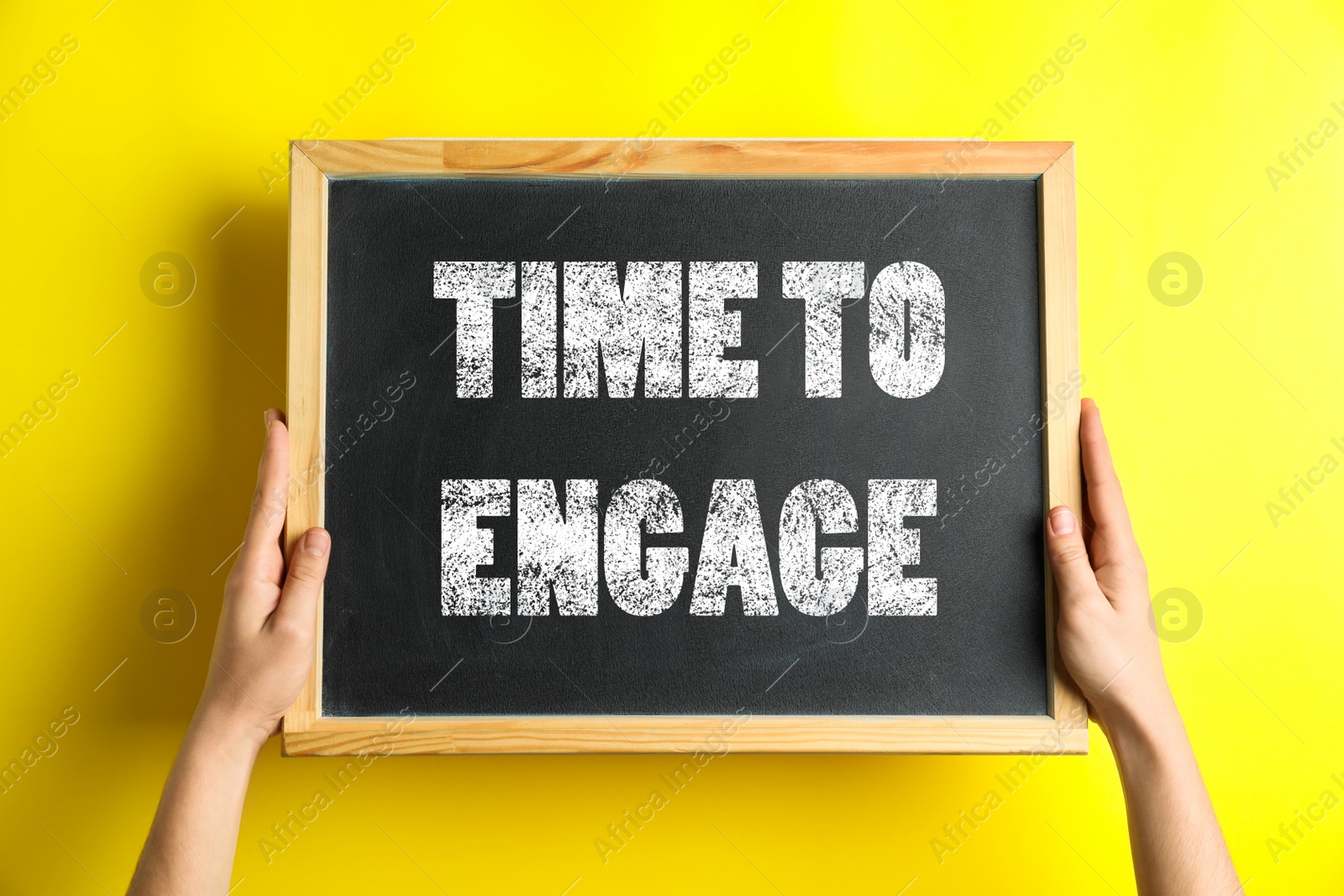 Image of Woman holding chalkboard with text Time To Engage on yellow background, closeup