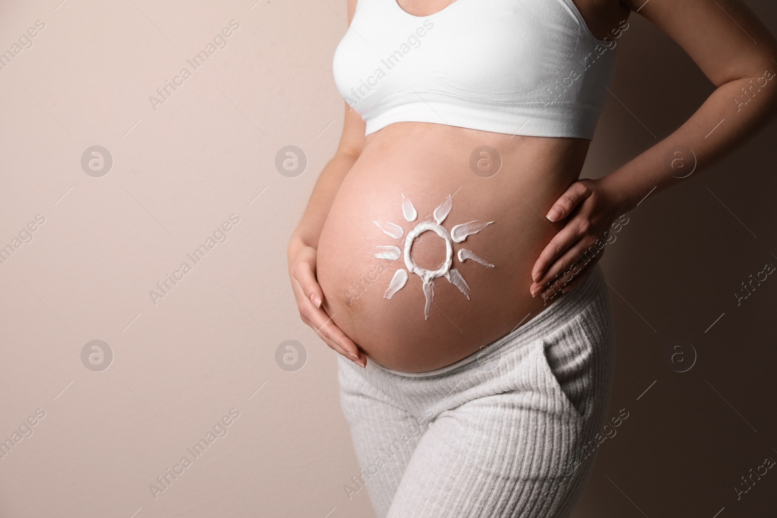 Photo of Young pregnant woman with sun protection cream on belly near beige wall, closeup. Space for text