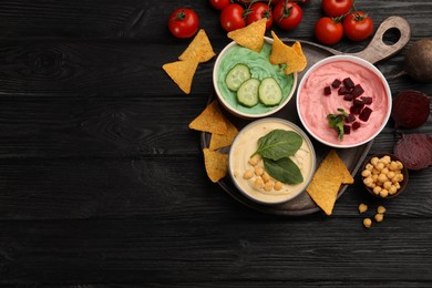 Photo of Different kinds of tasty hummus served with nachos and ingredients on black wooden table, flat lay. Space for text