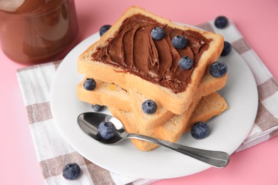 Photo of Tasty toast with chocolate paste and blueberries near jar on pink table, closeup