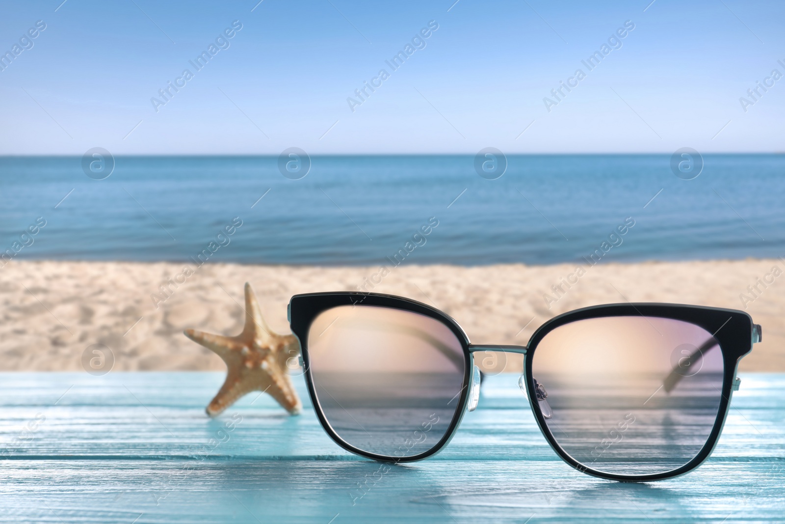 Image of Starfish and stylish sunglasses on light blue wooden table near sea with sandy beach