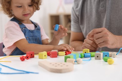Motor skills development. Father and daughter playing with wooden pieces and string for threading activity at table indoors, closeup