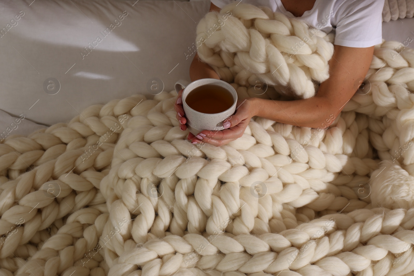 Photo of Woman covered with knitted plaid holding cup of tea in bed, closeup