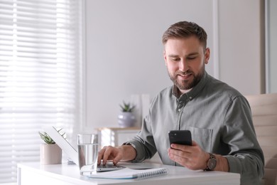 Young man with smartphone working on laptop at table in office