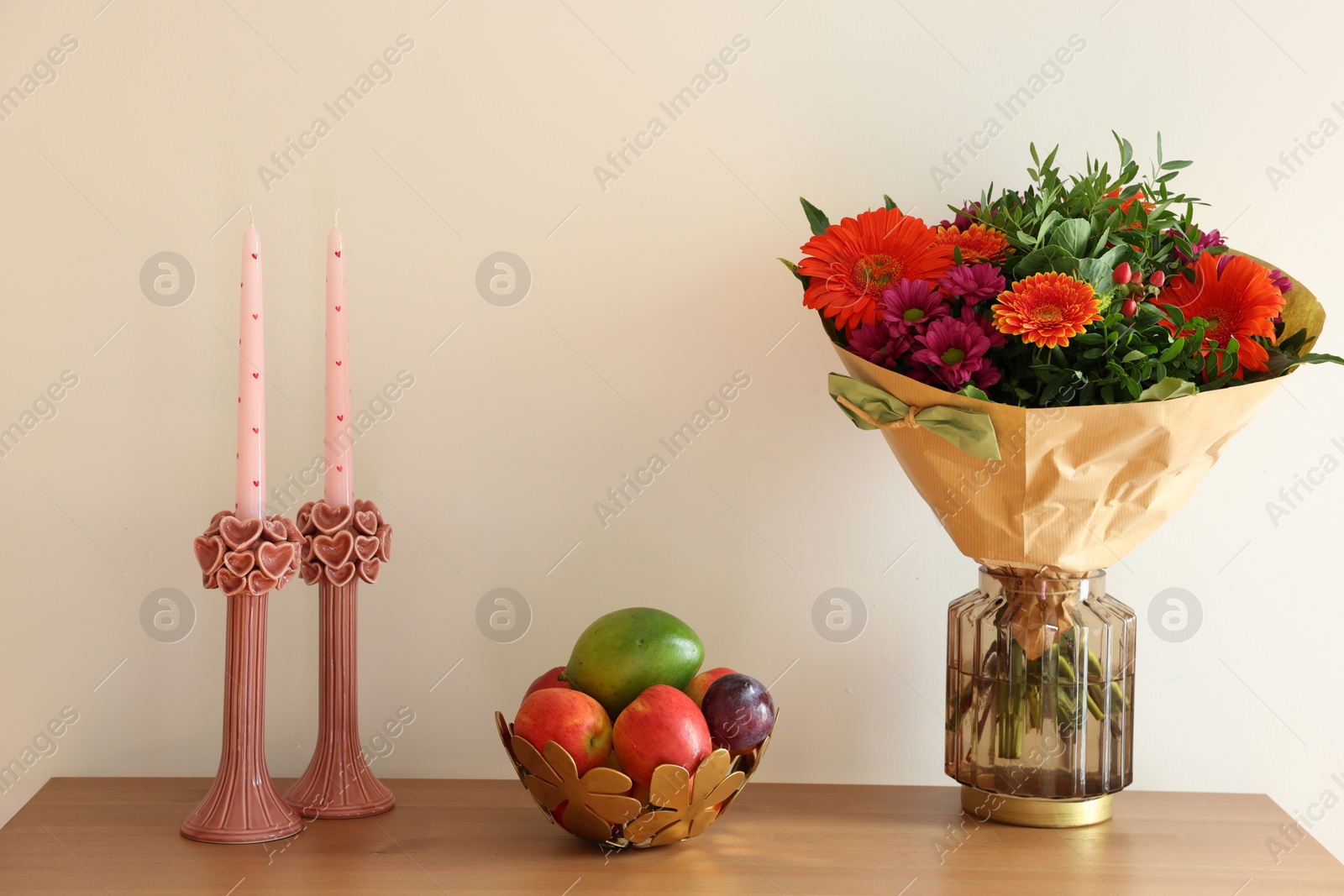 Photo of Bouquet of flowers, bowl with fresh fruits and candles on wooden table near white wall