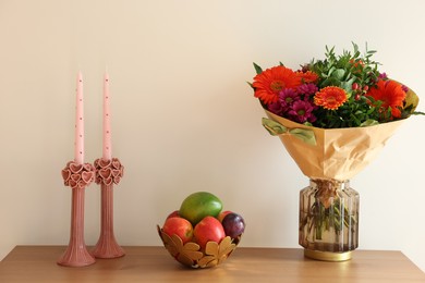 Photo of Bouquet of flowers, bowl with fresh fruits and candles on wooden table near white wall