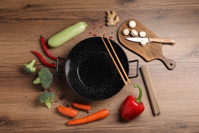 Empty iron wok surrounded by raw ingredients on wooden table, flat lay