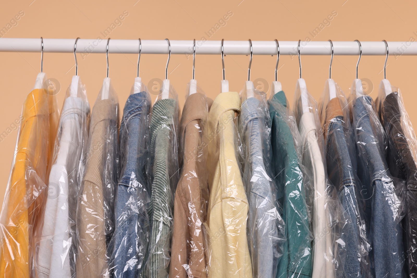 Photo of Dry-cleaning service. Many different clothes in plastic bags hanging on rack against beige background