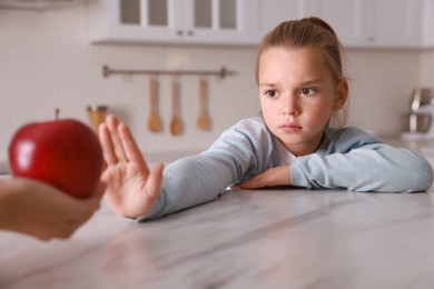 Photo of Cute little girl refusing to eat apple in kitchen, closeup