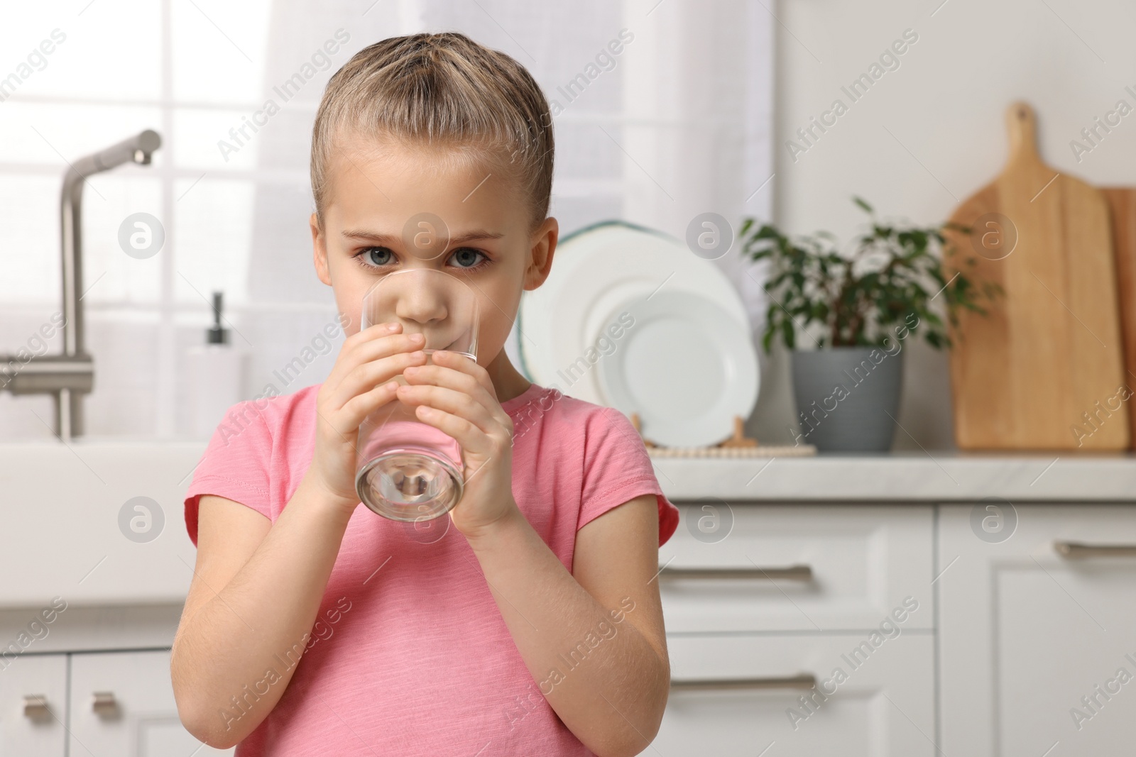 Photo of Cute little girl drinking fresh water from glass in kitchen