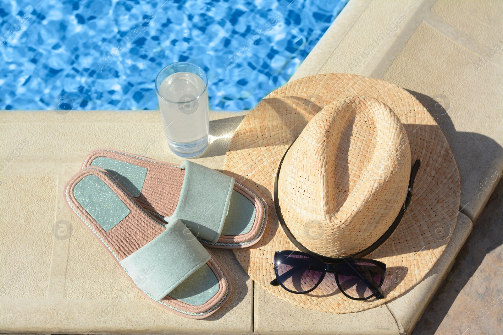 Photo of Stylish sunglasses, slippers, straw hat and glass of water at poolside on sunny day. Beach accessories