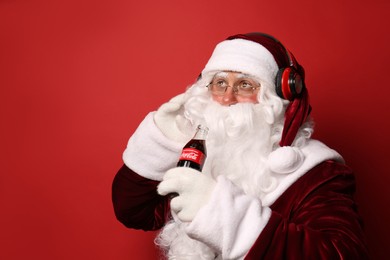 Photo of MYKOLAIV, UKRAINE - JANUARY 18, 2021: Santa Claus holding Coca-Cola bottle and listening to music with headphones on red background