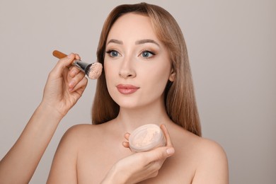 Professional makeup artist applying powder onto beautiful young woman's face with brush on grey background
