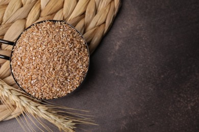 Photo of Dry wheat groats in scoop and spikelet on brown textured table, top view. Space for text