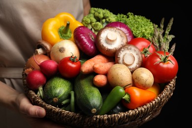 Photo of Woman holding wicker basket full of fresh vegetables on black background, closeup