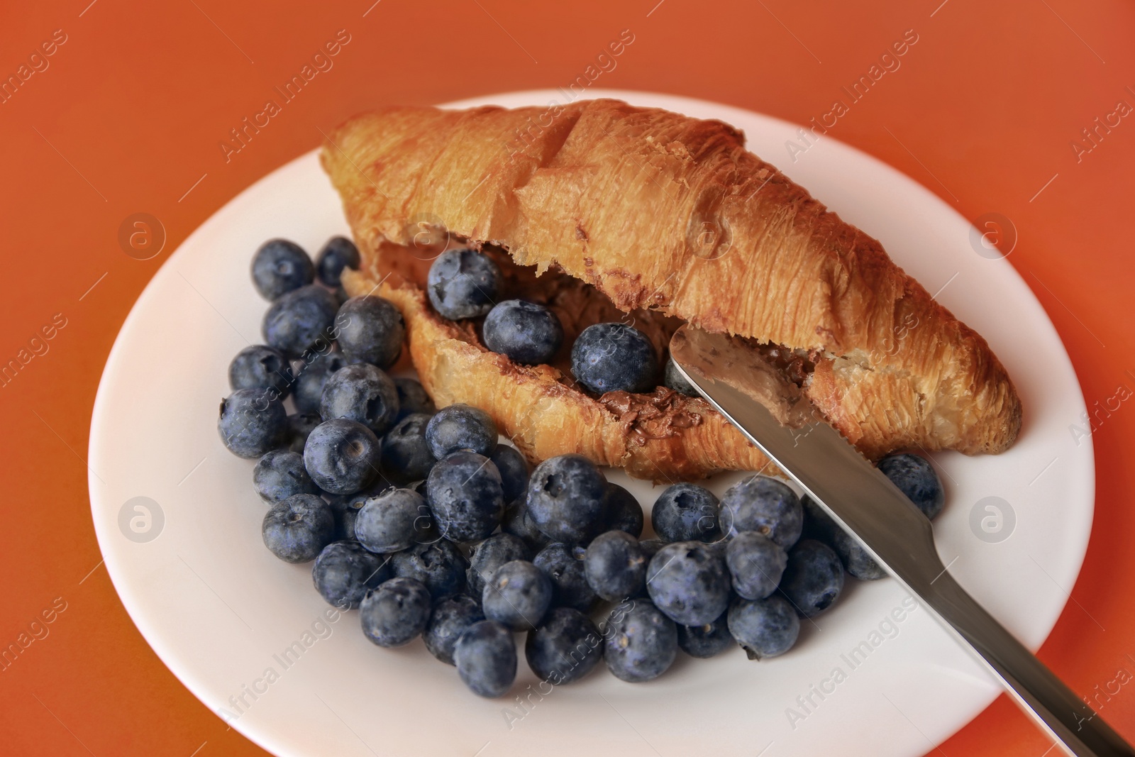 Photo of Tasty croissant with chocolate paste and blueberries on orange background, closeup