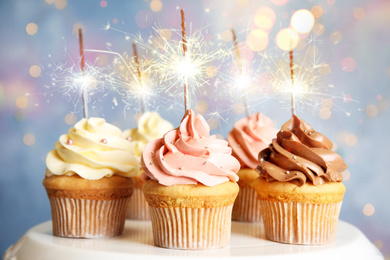 Delicious birthday cupcakes with sparklers on stand against blurred background