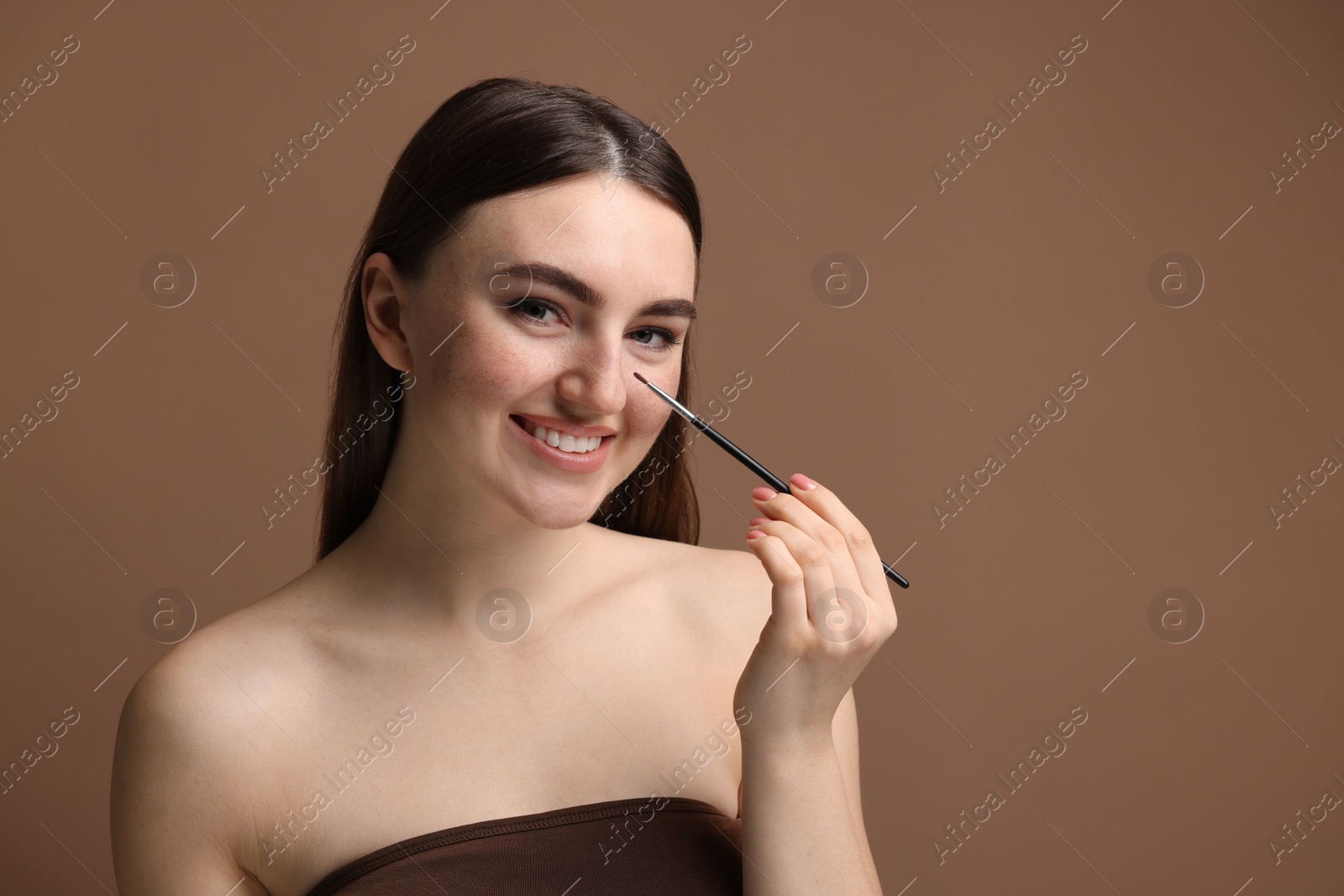 Photo of Smiling woman drawing freckles with brush on brown background