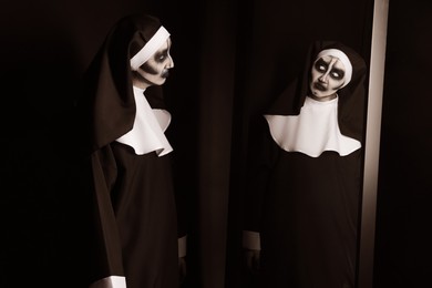 Photo of Scary devilish nun near mirror on black background. Halloween party look