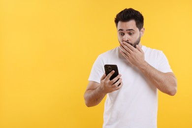 Shocked young man using smartphone on yellow background, space for text