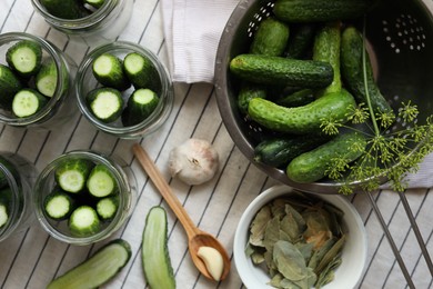 Photo of Glass jars with fresh cucumbers and other ingredients prepared for canning on tablecloth, flat lay