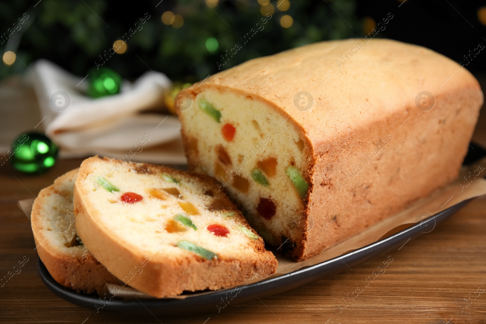 Photo of Delicious cake with candied fruits on wooden table against Christmas lights, closeup