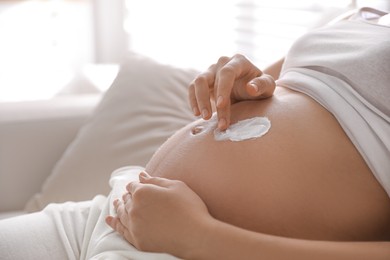 Photo of Young pregnant woman applying cosmetic product on belly indoors, closeup