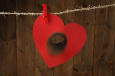 Photo of Red paper heart with burnt hole on rope near wooden wall. Broken heart