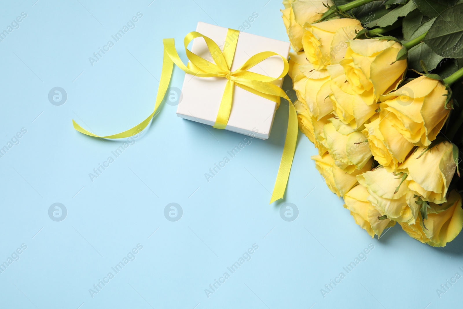 Photo of Beautiful bouquet of yellow roses and gift box on light blue background, flat lay. Space for text