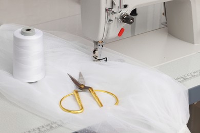 Photo of Sewing machine with white fabric, scissors and threads on table, closeup