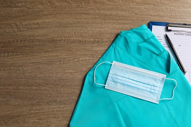 Medical uniform, protective mask and clipboard on wooden table, flat lay. Space for text