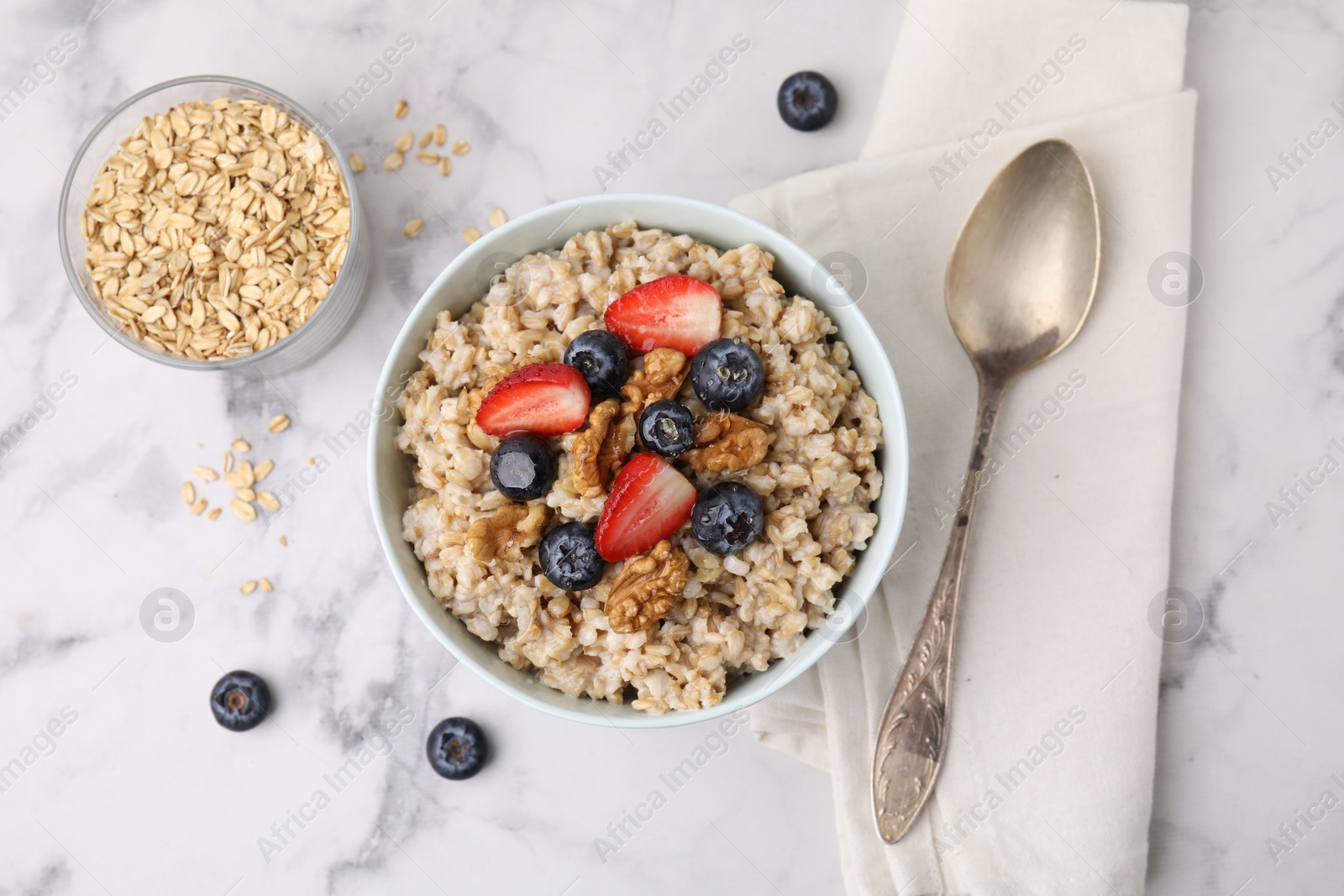 Photo of Tasty oatmeal with strawberries, blueberries and walnuts served on white marble table, flat lay