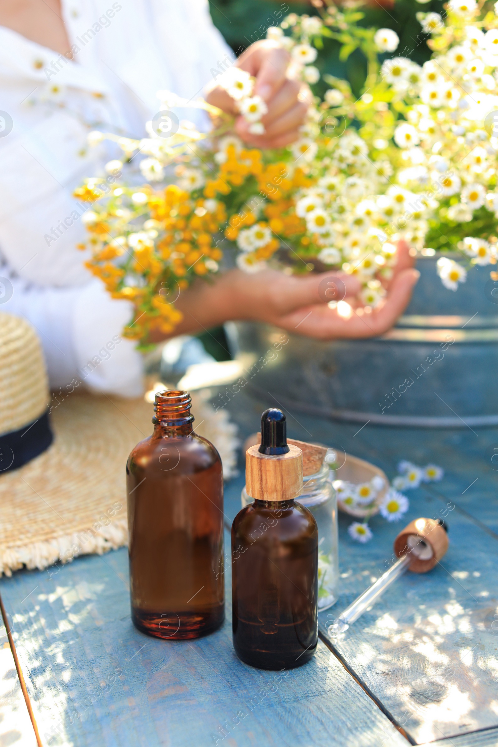 Photo of Woman with flowers near table outdoors, focus on bottles of chamomile essential oil