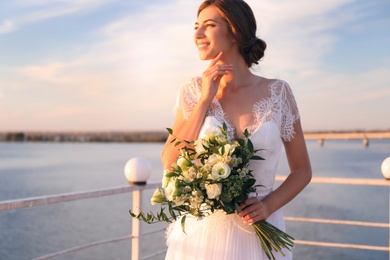 Photo of Gorgeous bride in beautiful wedding dress with bouquet near river, focus on flowers