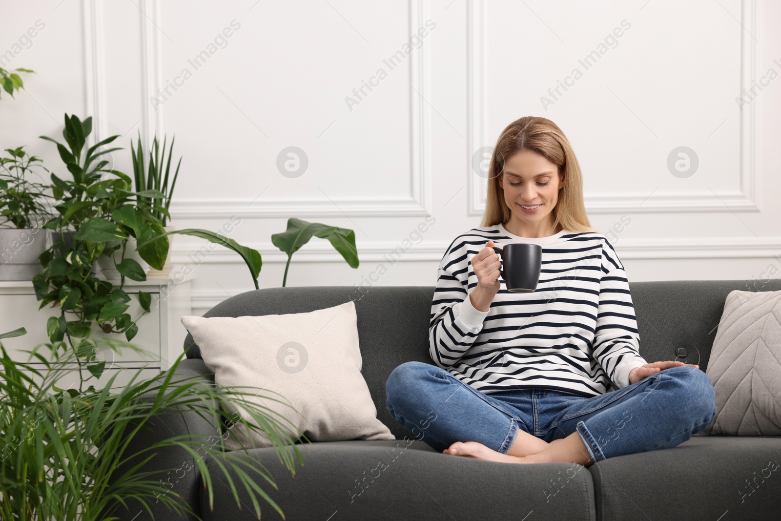 Photo of Woman with cup of drink sitting on sofa surrounded by beautiful potted houseplants at home