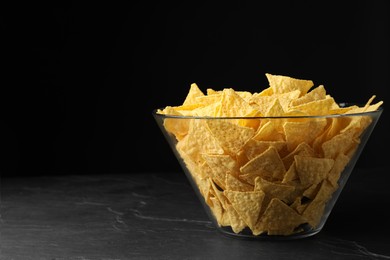 Photo of Glass bowl with tortilla chips (nachos) on black table against dark background, space for text