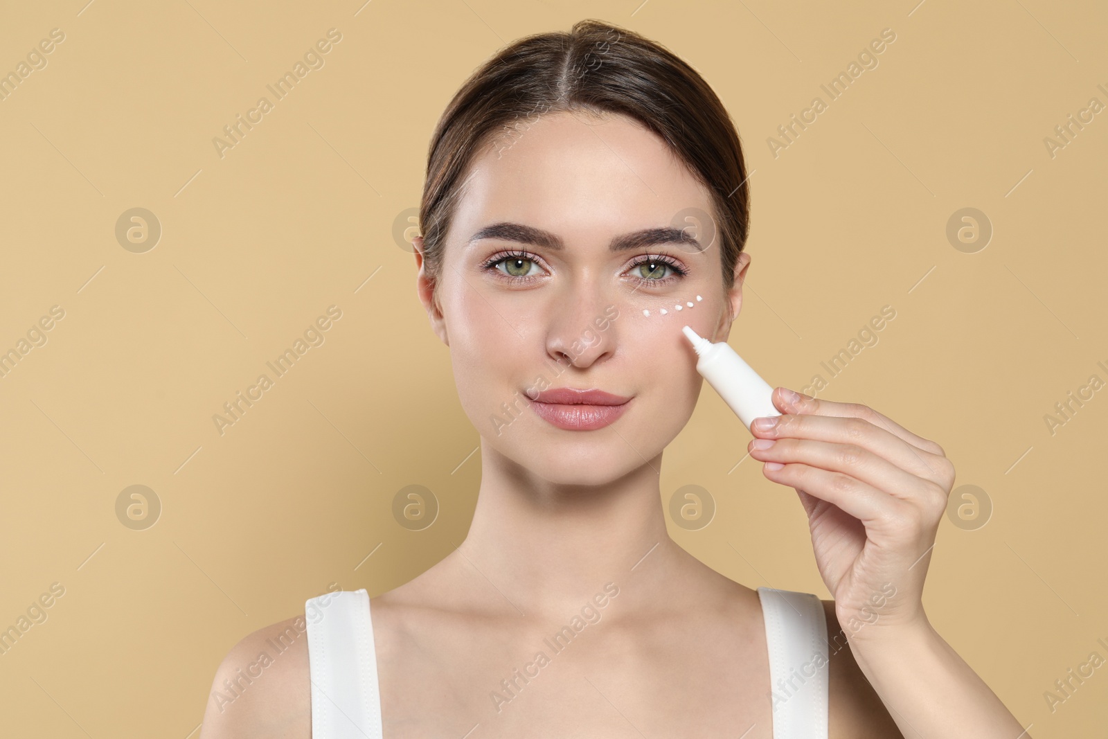 Photo of Young woman applying cream under eyes on beige background, space for text