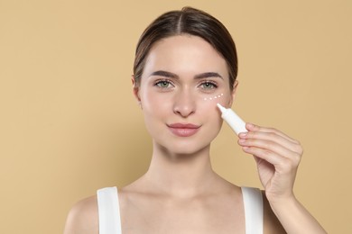 Young woman applying cream under eyes on beige background, space for text