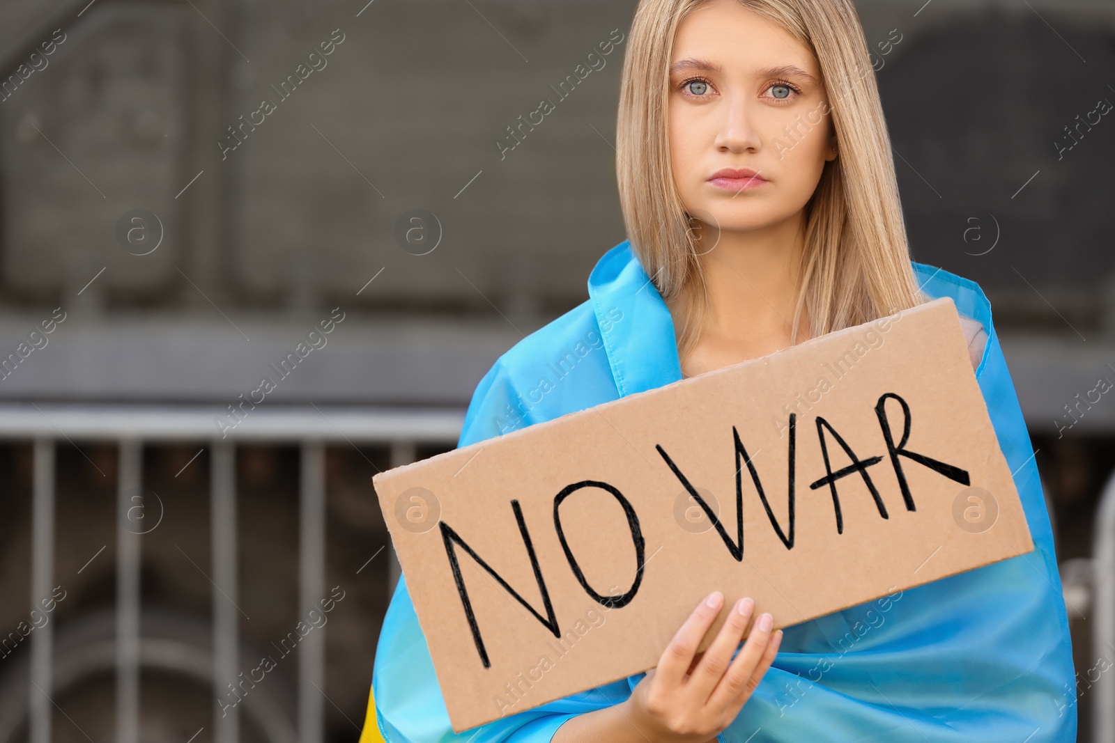 Photo of Sad woman wrapped in Ukrainian flag holding poster with words No War on city street