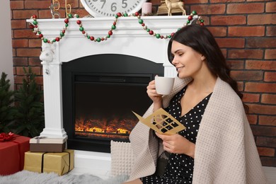 Young woman with greeting card and hot drink sitting near fireplace indoors