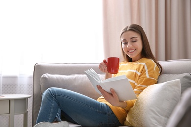 Photo of Young woman with cup of coffee reading book on sofa at home