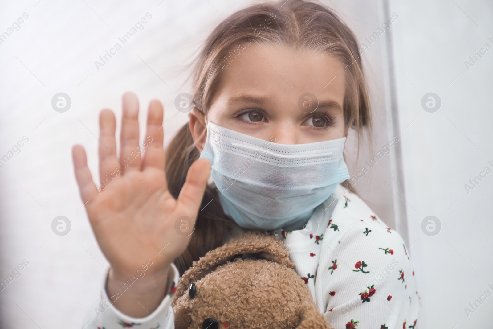 Photo of Sad little girl in protective mask looking out of window indoors, view from outside. Staying at home during coronavirus pandemic