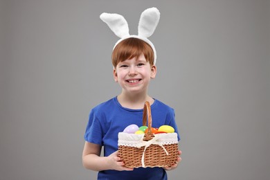 Easter celebration. Cute little boy with bunny ears and wicker basket full of painted eggs on grey background