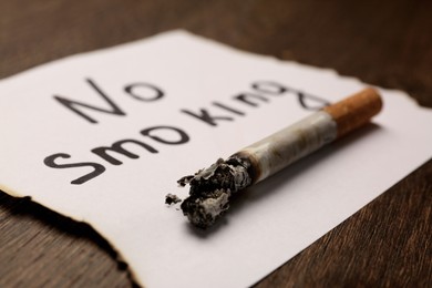 Burnt cigarette and words No Smoking written on paper on wooden table, closeup