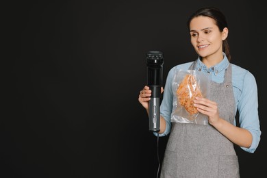 Beautiful young woman holding sous vide cooker and meat in vacuum pack on black background. Space for text