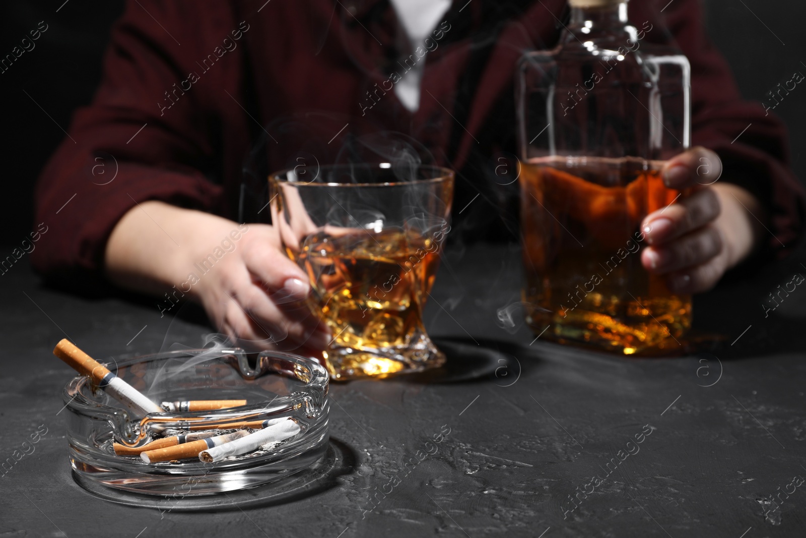 Photo of Alcohol addiction. Woman with whiskey and smoldering cigarettes at dark textured table, selective focus