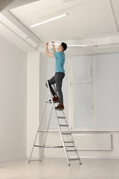 Photo of Young man installing ceiling lamp on stepladder indoors