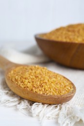 Napkin, spoon and bowl with uncooked bulgur on white wooden table, closeup